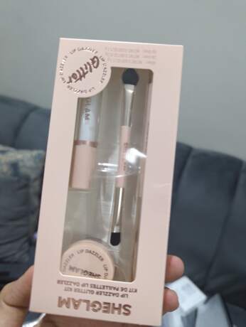 งบรรจุภัณฑ์สีชมพูอ่อนของชุด SHEGLAM Lip Glitter Kit ที่ถูกมือถืออยู่ แสดงให้เห็นองค์ประกอบภายในชุดผ่านช่องใส คือ ลิควิดลิปสติก กระปุกกลิตเตอร์ และแปรงสำหรับทา